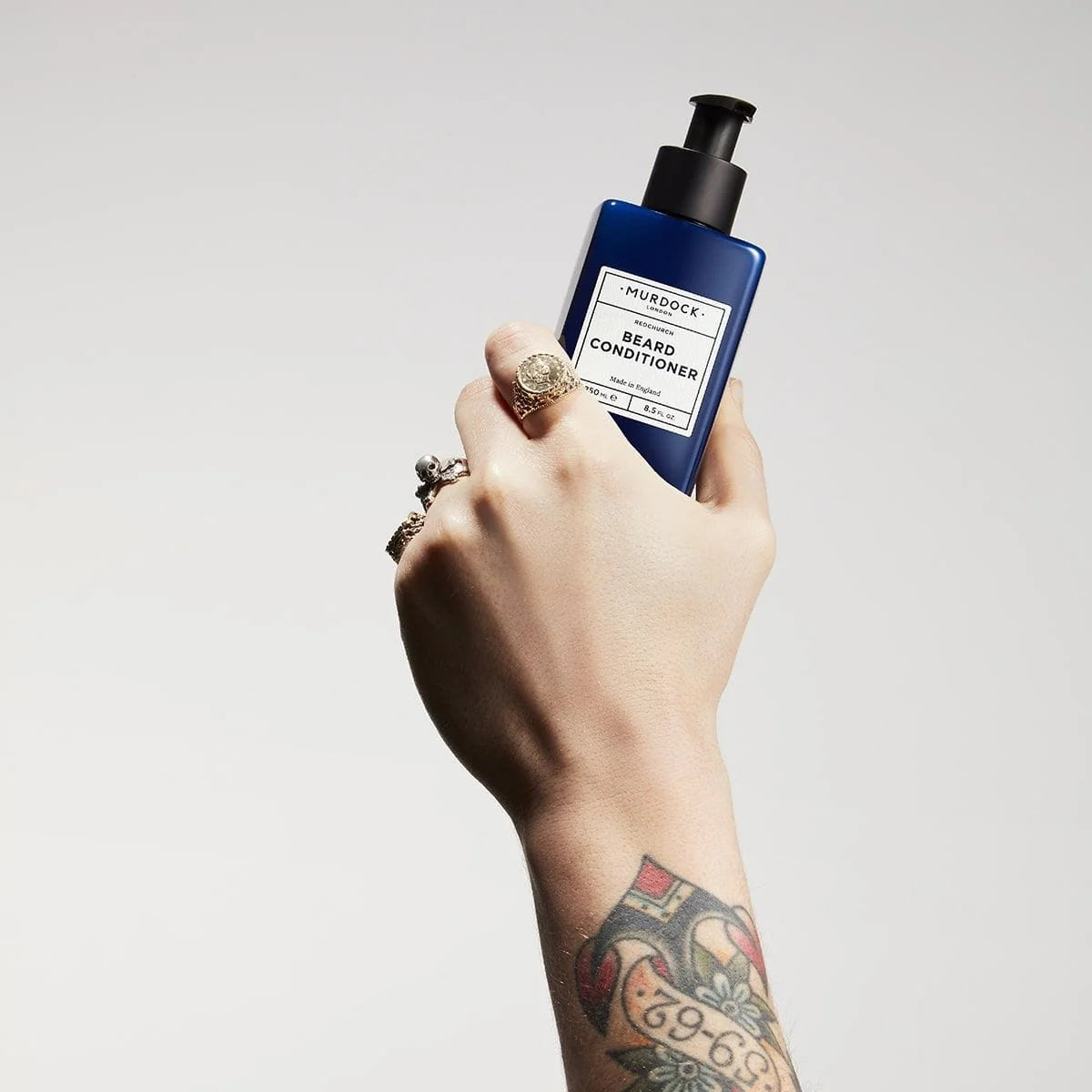 A hand with rings and a tattoo holds a blue bottle of Shueduck Beard Conditioner against a light gray background.